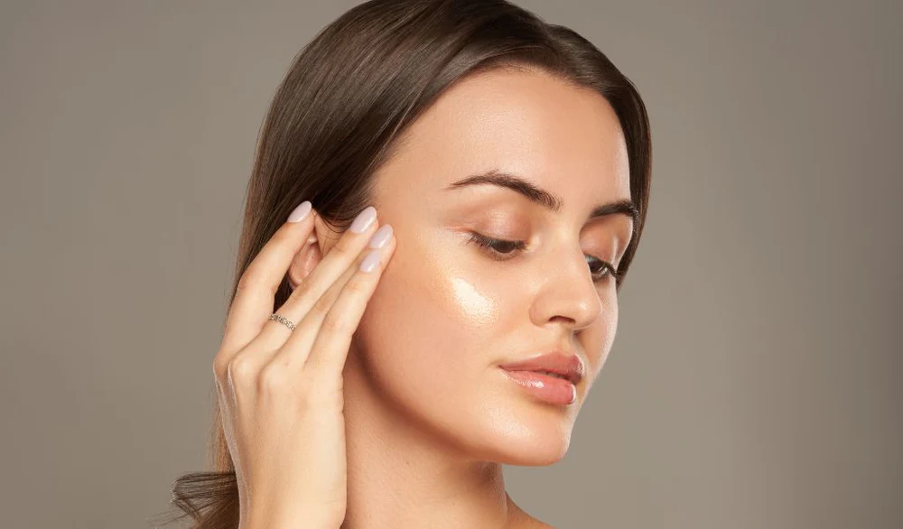 Woman with glowing skin and natural makeup gently touching her face, highlighting a smooth and radiant skincare look against a neutral background.